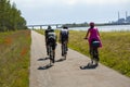 Three professional bicycle riders and one relaxed woman in pink dress cycling in nature by the sea with green grass and poppies on