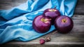 three purple apples sitting on top of a blue cloth next to a garlic plant and a blue cloth on a wooden table top with a blue Royalty Free Stock Photo