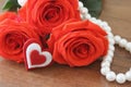 Three red roses closeup bouguet, a souvenir in the shape of a heart and a necklace of pearly white lying on a wooden table broown.