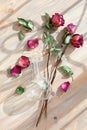 Three red roses, scattered flower petals, green leaves, glass round vase on wooden background top view closeup, floral arrangement