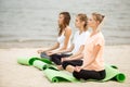 Three relaxed young girls sit in the lotus positions with closing eyes doing yoga on mats on sandy beach on a warm day