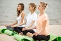 Three relaxed young girls sit in the lotus positions with closing eyes doing yoga on mats on sandy beach on a warm day