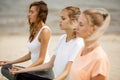 Three relaxed young girls sit in the lotus positions with closing eyes doing yoga on mats on sandy beach on a warm day