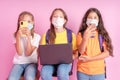 Three schoolgirls in medical masks with phones and a laptop in their hands are looking at the camera on a pink background.