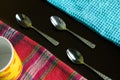 Three silver teaspoons between blue and multicolor napkins with yellow cup on the dark reflective backdrop.