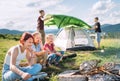 Three sisters cheerfully laughing and roasting marshmallows and candies on sticks over campfire flame while two brothers set up