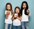 Three socked kids girls friends in white t-shirts and blue jeans stand holding glasses of water, milk and fresh juice