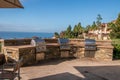 Three gas barbecue grills built into a stone wall on a patio overlooking the ocean and a resort