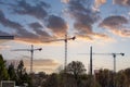 Three tall tower cranes surrounded by bare winter trees and lush green trees with blue sky and powerful clouds at sunset