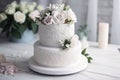 a three tiered white wedding cake with white flowers on top of a white table with a candle and flowers in the backround of the Royalty Free Stock Photo