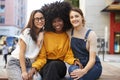 Three trendy young adult girlfriends on a bench in a city street smiling to camera, close up