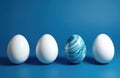 Three white eggs, one blue marbled egg stand in row on blue surface. Uniquely decorated egg stands out against plain ones. Royalty Free Stock Photo