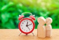 Three wooden human figures standing next to a red alarm clock on a nature background. Time concept. Business, Hourly payment