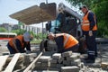 Three workers in orange vests using a mini backhoe loader mount a curb at the crossroads of city streets. May 29, 2020, Russia,
