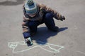 Three-year-old boy in colored outerwear and hat draws chalk on the asphalt in early spring