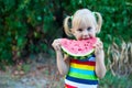 Three-year-old little European blonde girl eating a watermelon on a background of green leaves