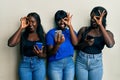 Three young african american friends using smartphone doing ok gesture with hand smiling, eye looking through fingers with happy Royalty Free Stock Photo