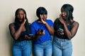 Three young african american friends using smartphone hand on mouth telling secret rumor, whispering malicious talk conversation Royalty Free Stock Photo