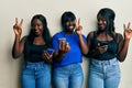 Three young african american friends using smartphone smiling with happy face winking at the camera doing victory sign with Royalty Free Stock Photo