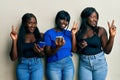 Three young african american friends using smartphone smiling looking to the camera showing fingers doing victory sign Royalty Free Stock Photo