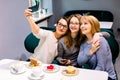 Three young beautiful women sitting at cafe indoors, happy, having fun, smiling, looking at smartphone, taking selfie