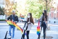 Three young caucasian and asian homosexual friends riding scooters with LGBT flags wearing face masks.