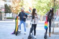 Three young caucasian and asian homosexual friends riding scooters with LGBT flags wearing face masks. LGBT pride celebration in