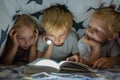 Three young children are reading a book with a flashlight under the covers at night. Royalty Free Stock Photo