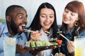 Three young excited and hungry multiethnic people, eating Japanese sea food sushi rolls at restaurant with happiness and