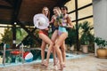 Three young female models posing in swimsuits holding pineapples, hat and juice at swimming pool at spa center