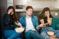 Three young friends two beautiful women and handsome man with remote control watching drama movie on the sofa eating pop corn,