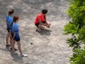 Three young girls holding petanque balls waiting to play on a sand floored petanque court. One girl squatting and the other two