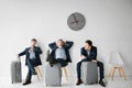 Three young men sit on white chairs and wait for flight. They look similar. Second hold legs on suitcase and lean to Royalty Free Stock Photo