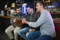 Three young men in casual clothes are smiling and clanging glasses of beer together while sitting at bar counter in pub Royalty Free Stock Photo