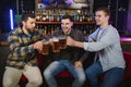 Three young men in casual clothes are smiling and clanging glasses of beer together while sitting at bar counter in pub Royalty Free Stock Photo