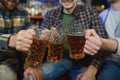 Three young men in casual clothes are smiling and clanging glasses of beer together while sitting at bar counter in pub Royalty Free Stock Photo