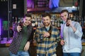 Three young men in casual clothes are smiling and clanging glasses of beer together while sitting at bar counter in pub Royalty Free Stock Photo