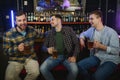 Three young men in casual clothes are smiling, holding bottles of beer while standing near bar counter in pub Royalty Free Stock Photo