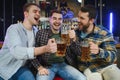 Three young men in casual clothes are smiling, holding bottles of beer while standing near bar counter in pub Royalty Free Stock Photo