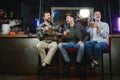 Three young men in casual clothes are smiling, holding bottles of beer while standing near bar counter in pub Royalty Free Stock Photo