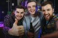 Three young men in casual clothes are smiling, holding bottles of beer while standing near bar counter in pub Royalty Free Stock Photo