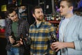 Three young men in casual clothes are smiling, holding bottles of beer while standing near bar counter in pub Royalty Free Stock Photo