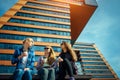 Three young pretty women sit on a bench on a city street, drinking coffee from disposable glasses, talking and laughing. Friendly Royalty Free Stock Photo