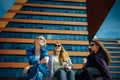 Three young pretty women sit on a bench on a city street, drinking coffee from disposable glasses, talking and laughing. Friends Royalty Free Stock Photo