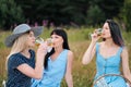 Three young women, blond, brunette and with dyed hair in blue dresses, and hats, sit on plaid and drink wine from Royalty Free Stock Photo