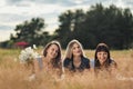 Three young women in blue dresses, and hats lie on plaid and drink wine. Outdoor picnic on grass on beach. Delicious