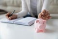 Thrifty woman writing daily expenses put coin into piggy bank Royalty Free Stock Photo