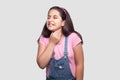 Throat or neck pain. Portrait of sick brunette young girl in pink t-shirt and blue overalls standing and holding her painful neck
