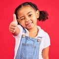 Thumbs up, portrait and girl child in studio, red background or isolated hand sign. Happy kids, thumb gesture and wink