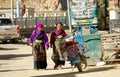 Two senior women in colorful traditional clothes walk down the unpaved street.